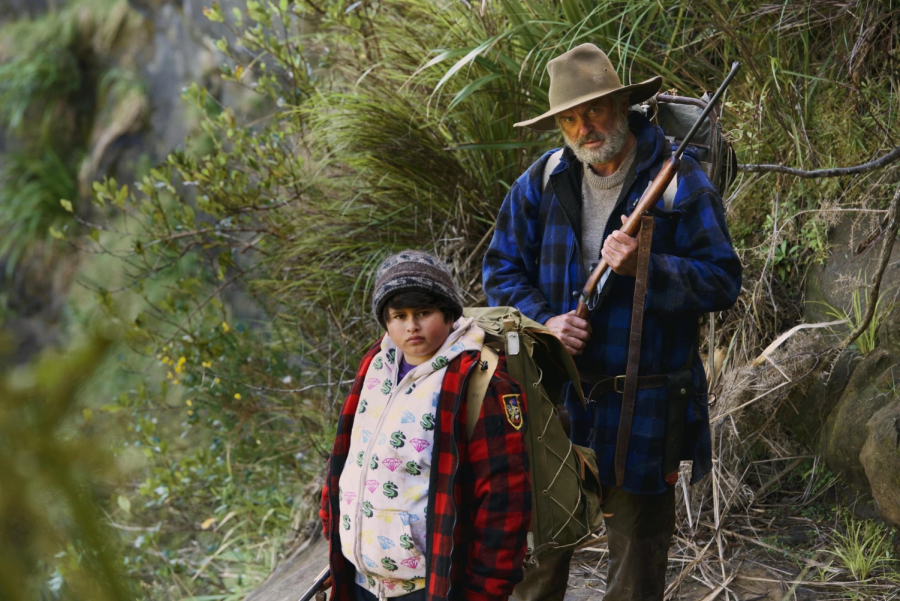 Picture of tall, older man and stout, younger boy staring at the camera. The younger boy is wearing a backpack, and the older man is holding a rifle. (Scene from Hunt for the Wilderpeople)